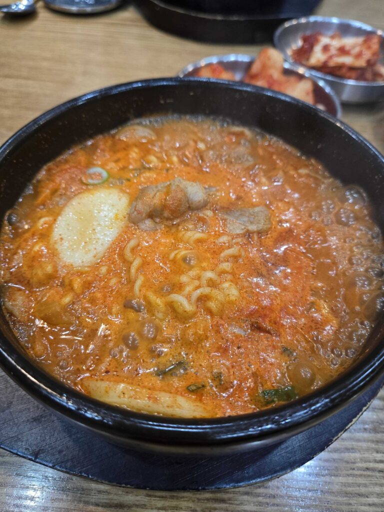 Budae suyuk gukbap close-up showing ramyeon noodles, sausages, and cheese in red broth