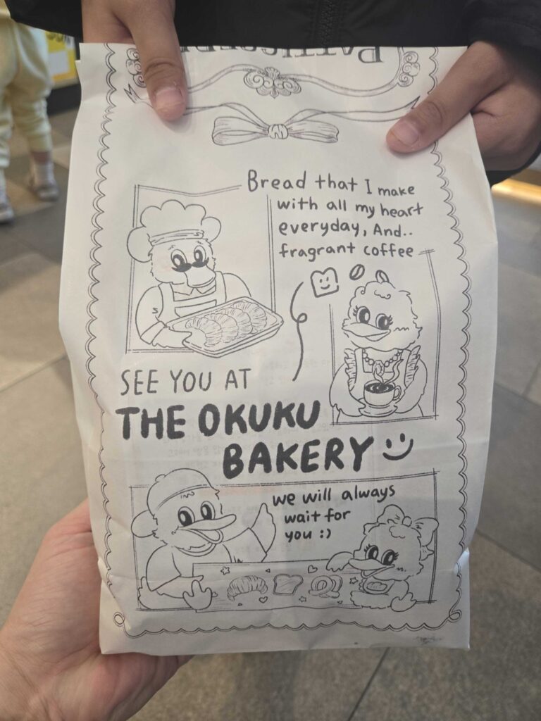 Okuku Bakery paper bag held in hand showing front illustration design with duck mascot characters