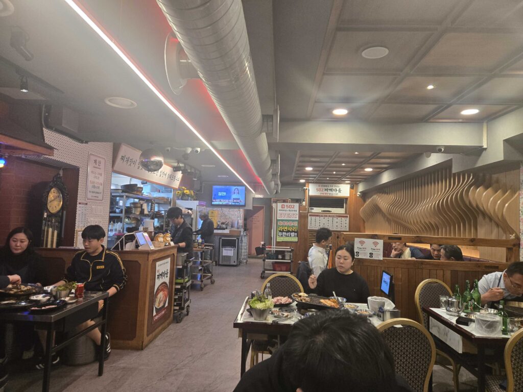 Busy interior of 502 Jjigae Maeul near Konkuk University showing customers dining at tables with open kitchen in background