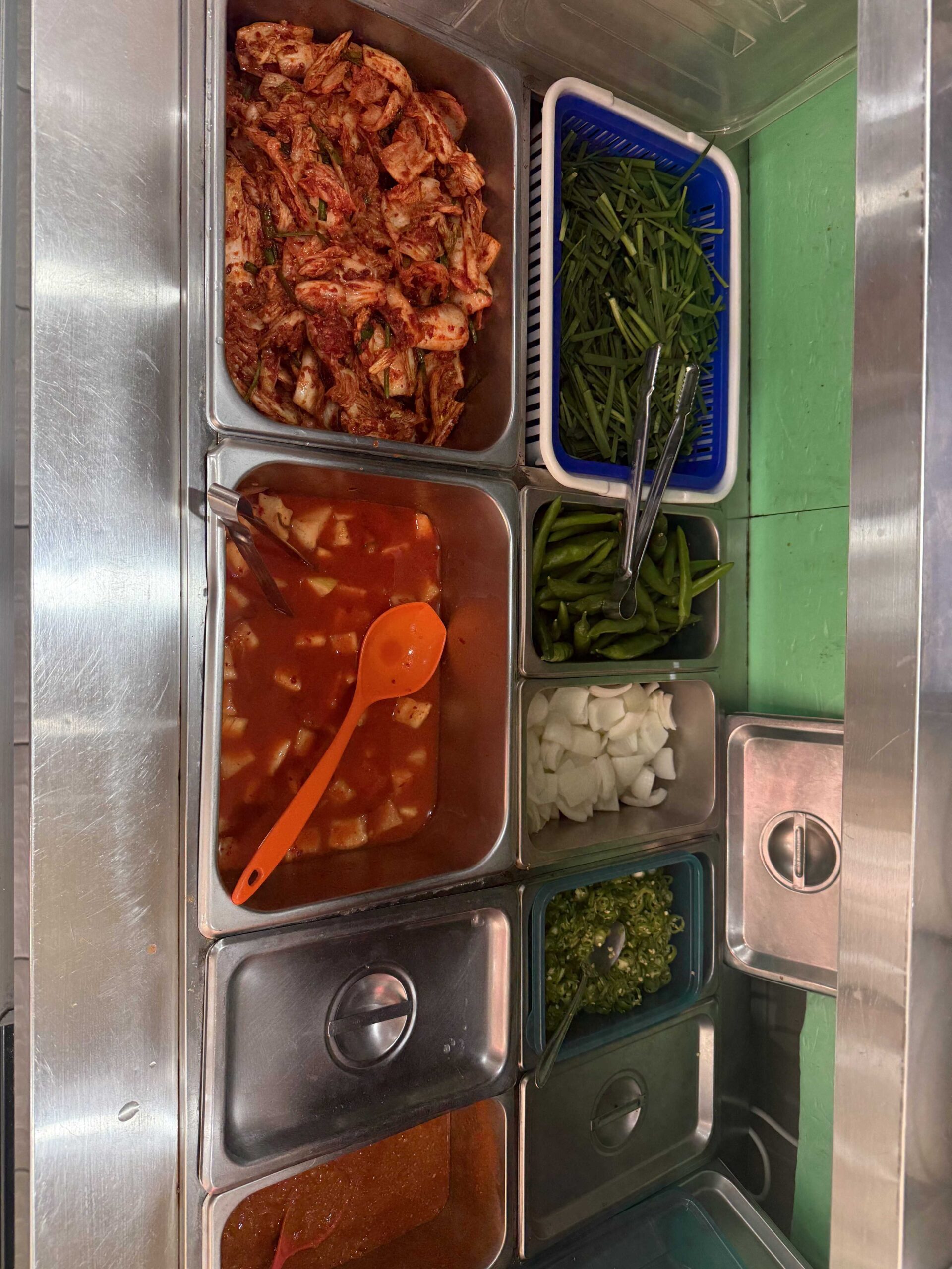 Self-serve banchan station with kimchi greens onion and chili at Dukkeobi Sundae Guk Hanam