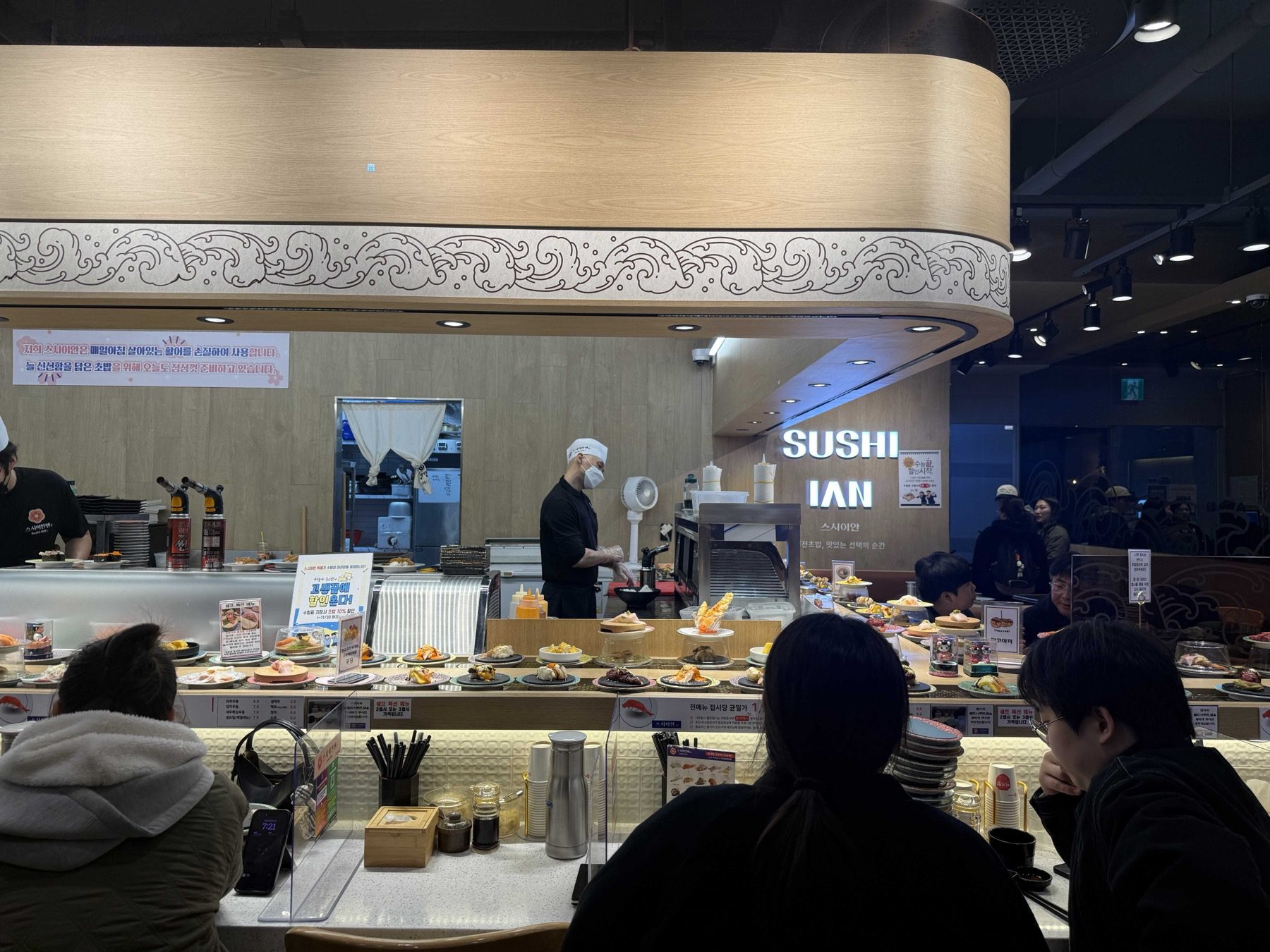 Sushi Ian Wirye counter with chef torching sushi and plates under clear domes on the belt