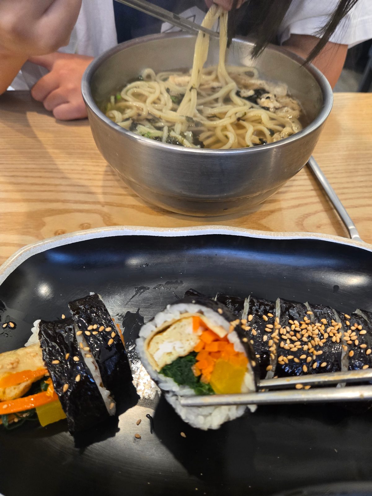 Child lifting udon noodles with chopsticks behind a plate of kimbap